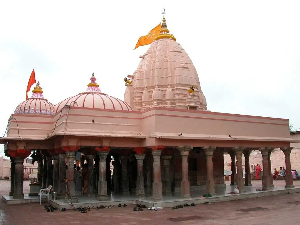 Exterior view of Gadkalika Temple in Ujjain, featuring traditional pink spires with golden accents, supported by ornate pillars, and adorned with saffron flags, with devotees gathered around.