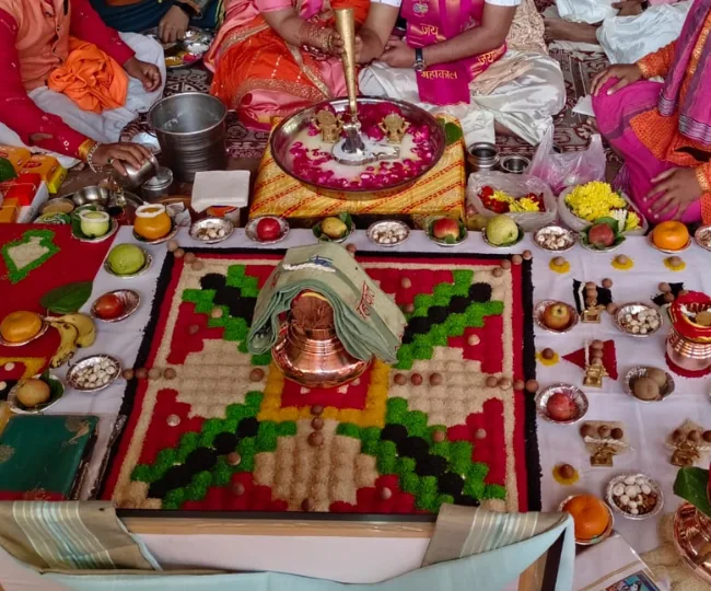 Traditional Mahamritunjay Jaap ritual setup with a colorful rangoli, copper pot, and offerings like fruits and flowers, surrounded by devotees in a spiritual ceremony.